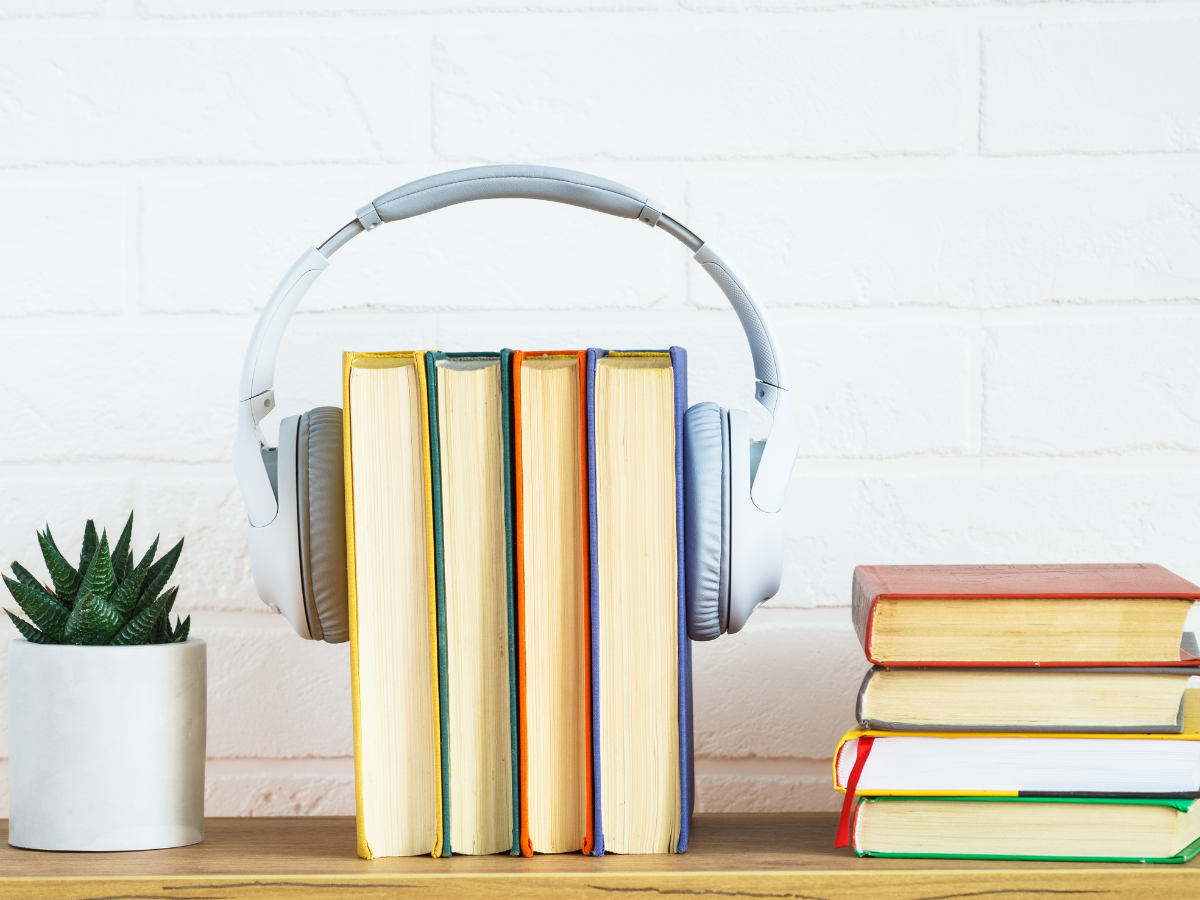 a row of books held together by over the ear headphones. a stack of books and a small potted plant sit on either side.