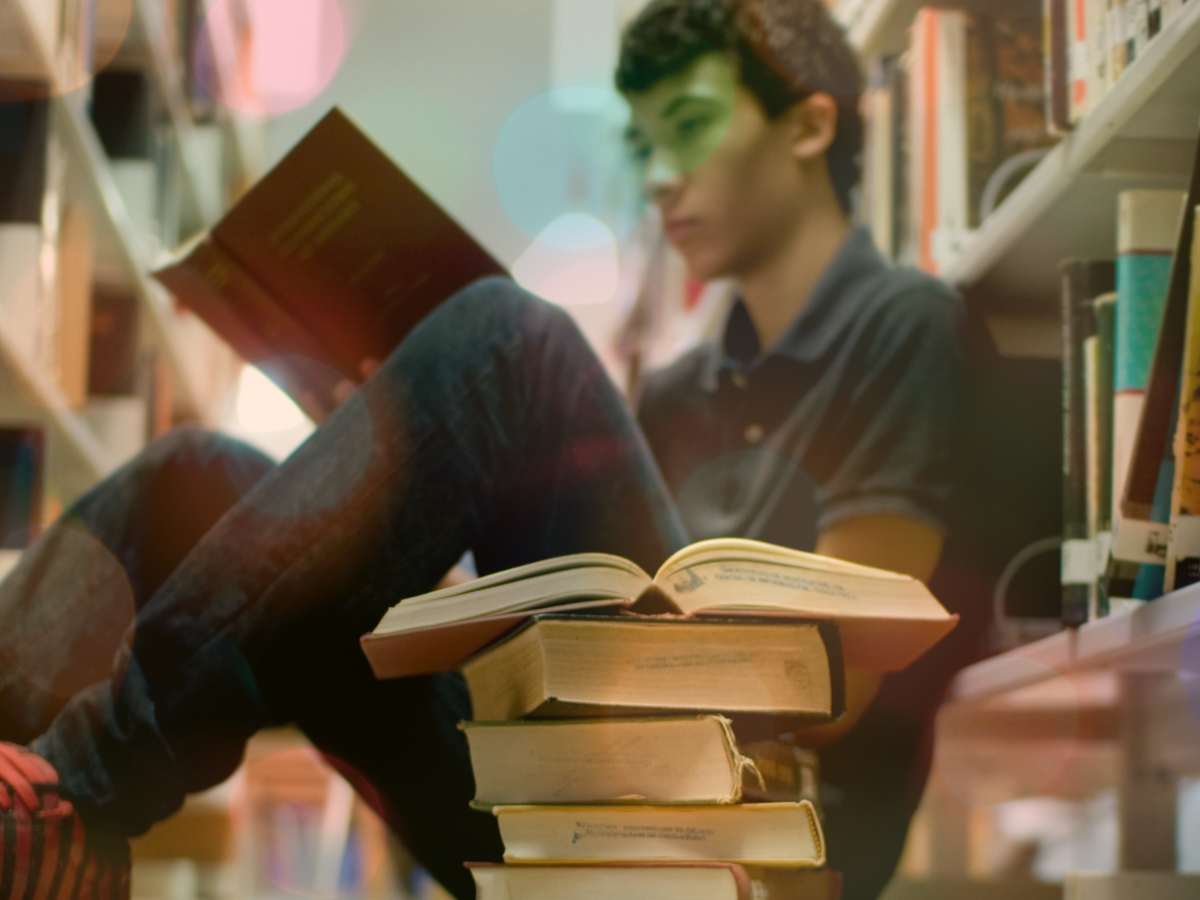 Teen sitting by a pile of books on a library floor reading a red book.