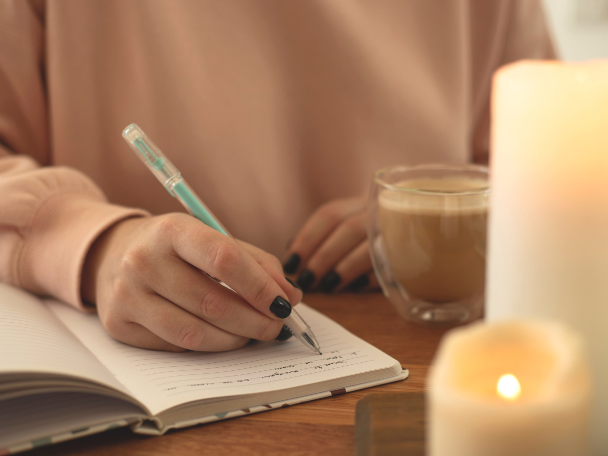 person in a pale pink sweater writing in a journal by a lit candle.